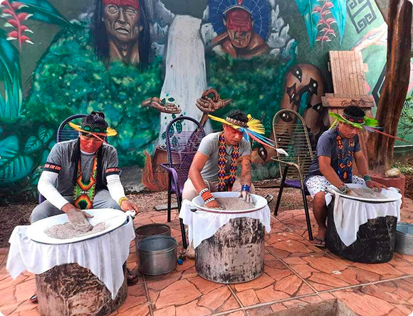 Three indigenous men in traditional attire preparing rapé in large containers, with a mural depicting ancestral figures in the background.